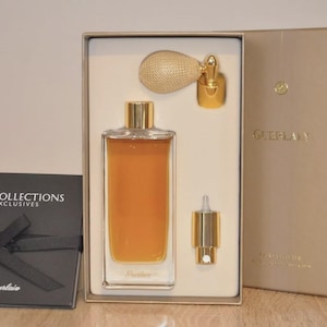 May include: A perfume set in a gold-toned box, featuring a rectangular glass bottle with amber liquid, a gold atomizer, and a small gold applicator. A black box with a ribbon and the text "LES COLLECTIONS EXCLUSIVES" is on the left.