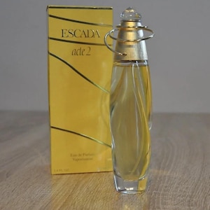 Puede incluir: Un frasco de perfume Escada Acte 2 de vidrio transparente y dorado. El frasco tiene una tapa dorada con un anillo alrededor. La caja es amarilla con el logotipo de Escada Acte 2 en negro.