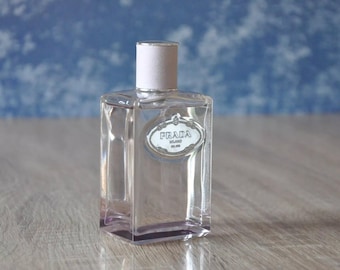 May include: A clear, rectangular Prada perfume bottle with a silver label and a light pink cap. The bottle is on a wooden surface with a blue background.