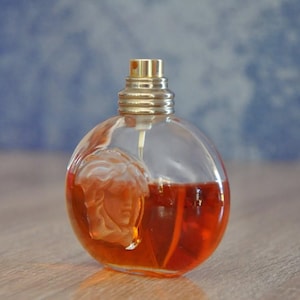 Pu&ograve; includere: Un flacone di profumo rotondo e trasparente con un tappo color oro e un disegno di volto scolpito. Il flacone &egrave; riempito con un liquido arancione-rossastro. Lo sfondo &egrave; blu sfocato e il flacone &egrave; su una superficie di legno.