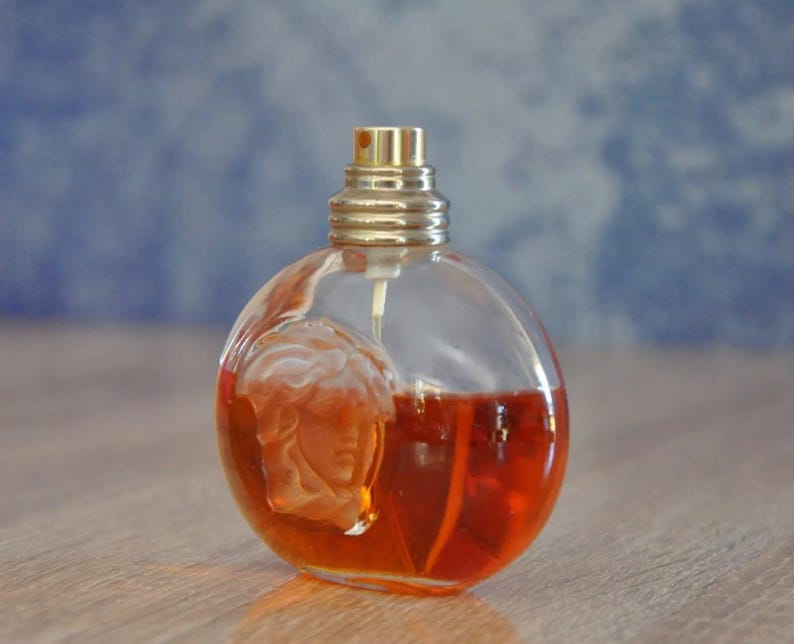 Pu&ograve; includere: Un flacone di profumo rotondo e trasparente con un tappo color oro e un disegno di volto scolpito. Il flacone &egrave; riempito con un liquido arancione-rossastro. Lo sfondo &egrave; blu sfocato e il flacone &egrave; su una superficie di legno.