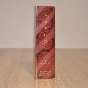 Puede incluir: Un frasco rojo y dorado de Trussardi Python Eau de Toilette Spray Natural. El frasco tiene un patrón de piel de serpiente y el texto "Python" está escrito verticalmente en letras doradas.
