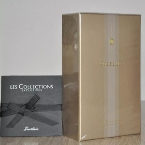 Puede incluir: Una caja dorada con el texto "Guerlain" en la parte delantera. La caja está sobre una superficie negra junto a una caja negra con el texto "Les Collections Exclusives Guerlain" en la parte delantera.