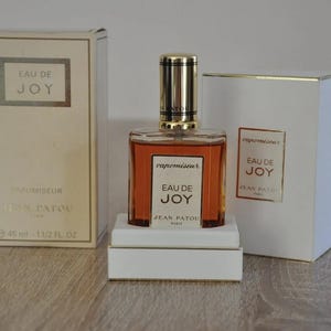Pode incluir: Um conjunto de três frascos de perfume com uma tampa dourada e um frasco de vidro transparente. Os frascos são etiquetados com "Eau de Joy" e "Jean Patou".