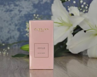 Könnte beinhalten: Eine rosa rechteckige Schachtel mit Guerlain Idylle Eau de Toilette. Die Schachtel hat goldfarbene Schrift und ein quadratisches Etikett mit dem Produktnamen. Weiße Lilien und ein blauer Hintergrund sind im Hintergrund.