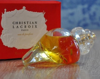 May include: A clear glass bottle of Christian Lacroix Paris eau de parfum. The bottle is shaped like a seashell and has a gold-colored liquid inside. The bottle is sitting on a wooden surface.