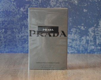 May include: A silver box with the word "PRADA" printed in black on the front. The text "AMBER POUR HOMME INTENSE" is printed in black on the bottom of the box.