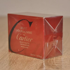 May include: A red Cartier Must de Cartier Pour Homme Eau de Toilette Vaporisateur Natural Spray 50ml 1.6 fl.oz. perfume box. The box is wrapped in clear plastic. The text on the box includes the brand name, product name, and size. The color scheme is primarily red with black and gold accents.