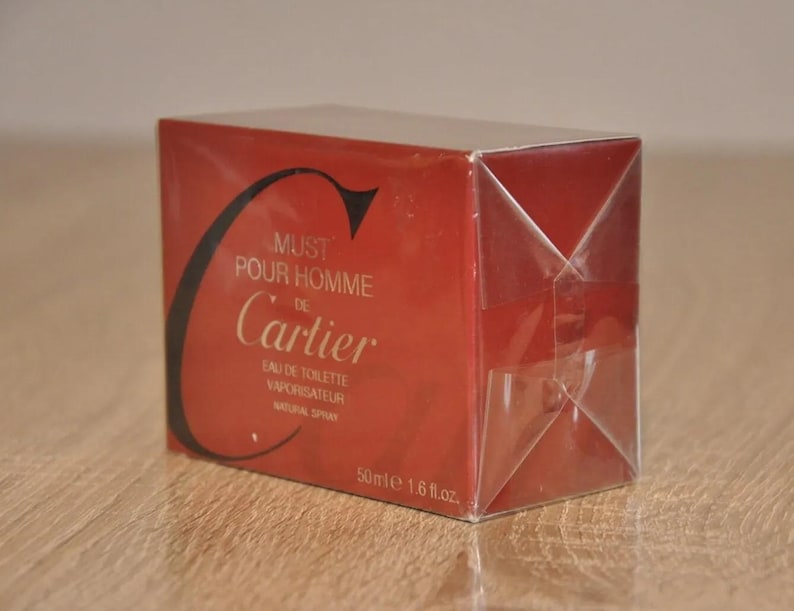 May include: A red Cartier Must de Cartier Pour Homme Eau de Toilette Vaporisateur Natural Spray 50ml 1.6 fl.oz. perfume box. The box is wrapped in clear plastic. The text on the box includes the brand name, product name, and size. The color scheme is primarily red with black and gold accents.