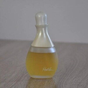 Può includere: Un flacone di profumo Halston in vetro trasparente con un tappo bianco. Il flacone ha la forma di una lacrima e ha un'etichetta dorata con la parola "Halston" scritta in corsivo.