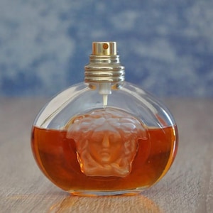 Pu&ograve; includere: Un flacone di profumo rotondo con tappo color oro e corpo in vetro trasparente. Il flacone contiene un liquido marrone rossiccio e presenta un disegno a testa di Medusa. Il flacone &egrave; appoggiato su una superficie di legno.