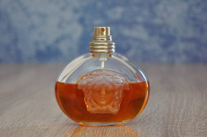 Pu&ograve; includere: Un flacone di profumo rotondo con tappo color oro e corpo in vetro trasparente. Il flacone contiene un liquido marrone rossiccio e presenta un disegno a testa di Medusa. Il flacone &egrave; appoggiato su una superficie di legno.