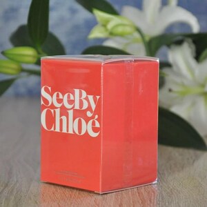 May include: A rectangular, coral-colored box with the words "See By Chlo&eacute;" in white lettering. The box is in front of white lilies and green foliage. The box has a clear plastic top.