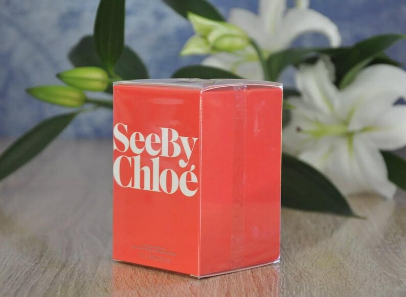 May include: A rectangular, coral-colored box with the words "See By Chlo&eacute;" in white lettering. The box is in front of white lilies and green foliage. The box has a clear plastic top.
