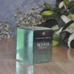 以下が含まれることがあります： ダイヤモンドのアイコンとTurquoise Diamond Wish Chopard Eau de Toilette Natural Spray 1.0 FL. OZ. 30 mlの文字が前面に印刷された、長方形のターコイズブルーのメタリックな香水箱。箱は反射する表面と鋭いエッジを持っています。
