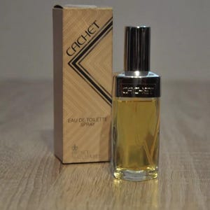 Può includere: Un flacone di vetro trasparente di spray di Eau de Toilette Cachet con un tappo dorato e un'etichetta dorata. Il flacone è su una superficie di legno. L'etichetta dice "Cachet Eau de Toilette Spray" e "Prince Matchabel".