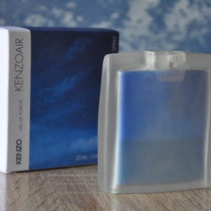 Puede incluir: Un juego de botella y caja de perfume. La botella es de forma rectangular esmerilada con un interior azul. La caja es azul oscuro con un diseño de nubes y el texto "KENZOAIR". El conjunto está sobre una superficie de madera.
