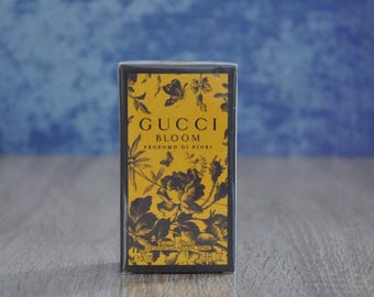 May include: A rectangular perfume box with a yellow and black floral design. The box features the text "GUCCI BLOOM PROFUMO DI FIORI" and contains 1.6 fl oz of perfume. The box has a black border and is set against a blue background.