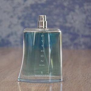 Puede incluir: Una botella de colonia de vidrio transparente con una tapa plateada. La botella contiene un líquido azul y la palabra "PYTHON" está impresa en blanco en la parte delantera.