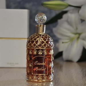 Könnte beinhalten: Eine goldene und braune Glasflasche mit Parfüm, einem goldenen Verschluss und einem Bienenmuster. Die Flasche ist mit "Oriental Brulant" beschriftet.