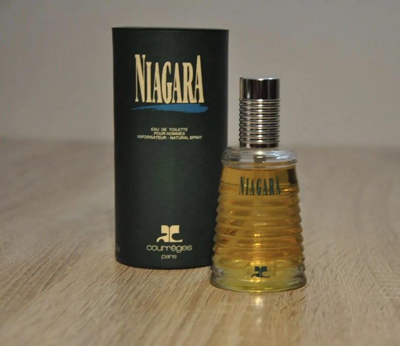 May include: A black and gold cylindrical bottle of Niagara cologne by Courr&egrave;ges Paris. The bottle has a textured design and a silver cap. The bottle is sitting on a wooden surface.