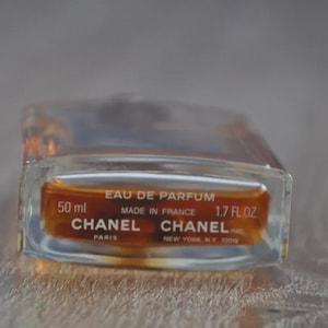 May include: A rectangular glass perfume bottle with a clear body and amber-colored liquid. The label reads "EAU DE PARFUM" and "CHANEL" with the volume of 50 ml and 1.7 fl oz. The bottle is made in France.