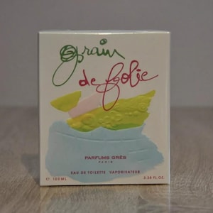 Op de afbeelding: Een witte doos met een abstract ontwerp in blauw, groen en geel. De tekst "Grain de folie" is geschreven in rood en groen. De doos is gelabeld met "Parfums Gres Paris" en "Eau de Toilette Vaporisateur". De doos is 100 ml of 3,38 fl oz.