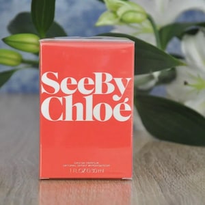 May include: A rectangular, coral-colored perfume box with the words "See By Chlo&eacute;" in large, white letters. The box also has smaller text indicating it is Eau de Parfum, 1 fl oz / 30 ml. White lilies and green leaves are in the background.