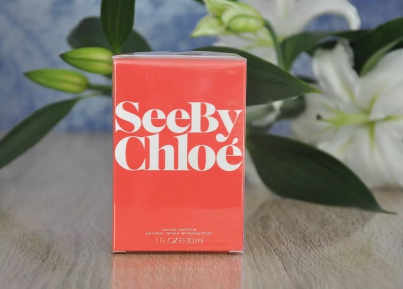 May include: A rectangular, coral-colored perfume box with the words "See By Chlo&eacute;" in large, white letters. The box also has smaller text indicating it is Eau de Parfum, 1 fl oz / 30 ml. White lilies and green leaves are in the background.