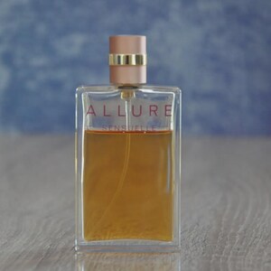 May include: A rectangular perfume bottle with the words "ALLURE SENSUELLE" in red lettering. The bottle contains a light amber liquid and has a light pink cap with a gold band. The background is a blurred blue and the bottle sits on a wooden surface.