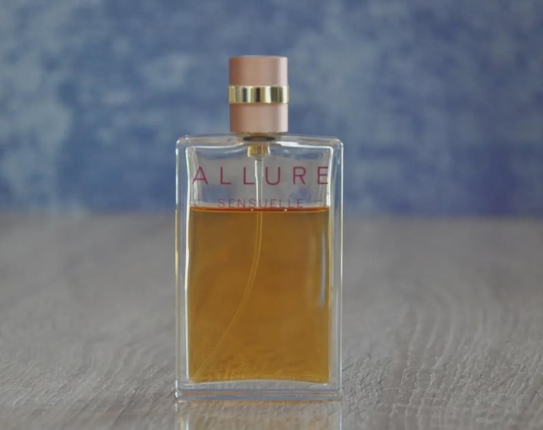 May include: A rectangular perfume bottle with the words "ALLURE SENSUELLE" in red lettering. The bottle contains a light amber liquid and has a light pink cap with a gold band. The background is a blurred blue and the bottle sits on a wooden surface.