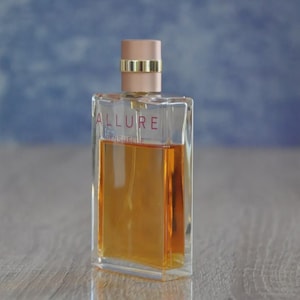May include: A rectangular perfume bottle with a clear glass body and a peach-colored cap. The bottle contains a golden-amber liquid and features the word "ALLURE" in red lettering. A gold band encircles the cap.