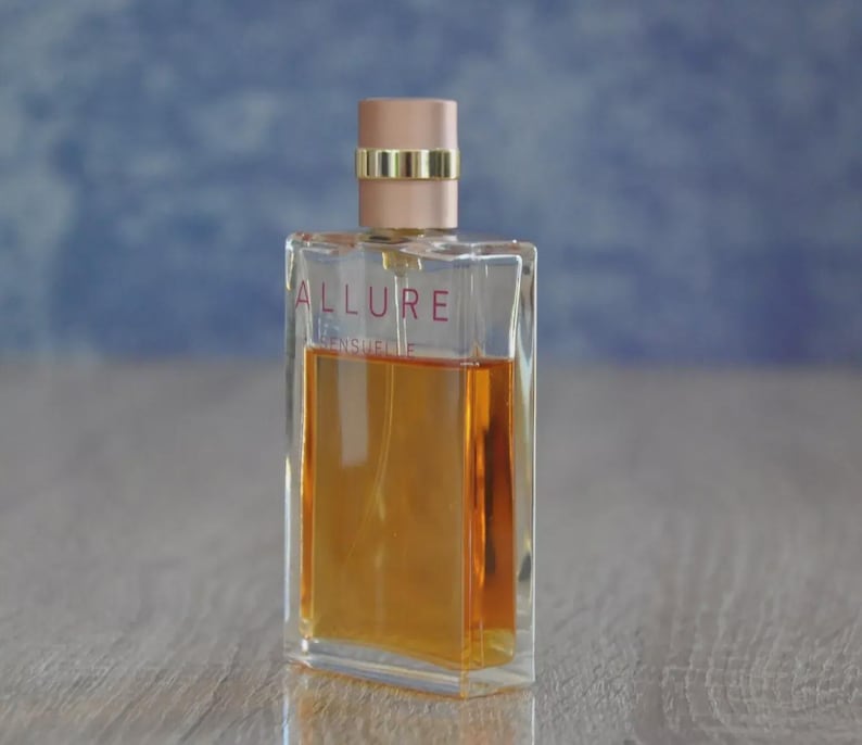 May include: A rectangular perfume bottle with a clear glass body and a peach-colored cap. The bottle contains a golden-amber liquid and features the word "ALLURE" in red lettering. A gold band encircles the cap.
