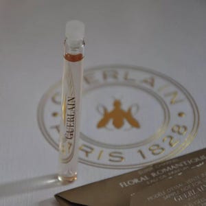 May include: A clear glass vial of Guerlain Floral Romantique Eau de Parfum. The vial has a white cap and contains a light amber liquid. The Guerlain logo and text are visible on the vial. A gold-colored Guerlain logo and text are in the background.