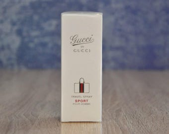May include: White travel spray bottle with a black and red Gucci logo and the text "Gucci by Gucci" and "Travel Spray Sport Pour Homme".