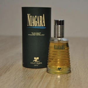 May include: A black and gold cylindrical bottle of Niagara cologne by Courr&egrave;ges Paris. The bottle has a textured design and a silver cap. The bottle is sitting on a wooden surface.