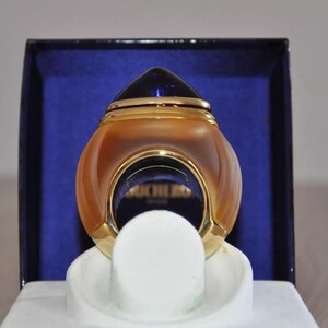 Op de afbeelding: Een gouden en blauwe parfumfles met een rond, ringvormig ontwerp. De fles staat op een wit voetstuk in een blauwe doos.
