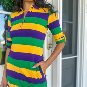 May include: A striped dress with green, yellow, and purple stripes. The dress has a collar and a button-up front. It has a pocket on the left side.