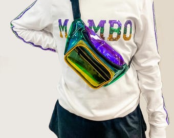 Può includere: Una maglietta bianca a maniche lunghe con la parola "MAMBO" in lettere di paillettes sul davanti. La maglietta ha un dettaglio a righe bianche e nere sulle maniche. Una marsupio arcobaleno iridescente è indossata sopra la maglietta.