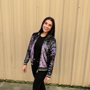 Puede incluir: Una mujer con una chaqueta bomber negra de lentejuelas con rayas moradas, verdes y doradas en los puños y el cuello. También lleva pantalones vaqueros negros y zapatos negros.