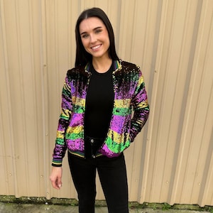 May include: A purple, green, and gold sequined bomber jacket with a black zipper and black ribbed trim. The jacket is worn over a black long-sleeved shirt and black jeans.