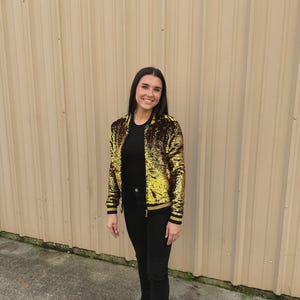 May include: A gold sequin bomber jacket with black and gold striped trim. The jacket is paired with black pants and a black top. The outfit is worn by a person standing in front of a corrugated metal wall.