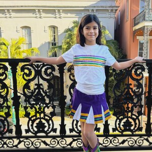 May include: A young person wearing a white t-shirt with a purple, green, and gold sequin stripe across the chest. They are also wearing a purple and white pleated skirt with a green and gold trim. The person is standing in front of a black wrought iron railing and wearing gold and green cowboy boots.
