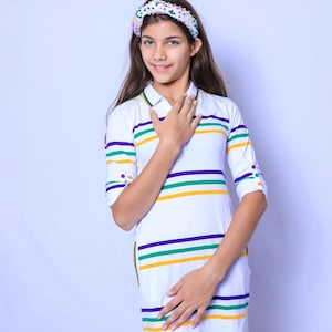May include: A young person wearing a white and colorful striped dress with a white headband with colorful beads. The dress has a collared neckline and three-quarter sleeves.