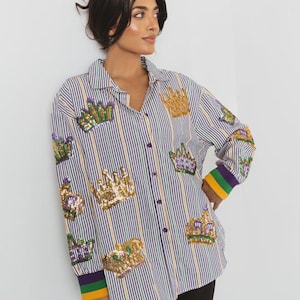Puede incluir: Camisa a rayas verticales en morado, blanco y amarillo, adornada con aplicaciones de coronas doradas y verdes con lentejuelas. Los puños presentan un diseño de bloques de color en morado, verde y amarillo. Cierre de botones.