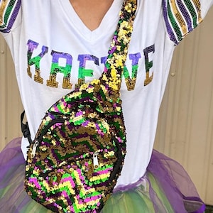 May include: A white long-sleeved shirt with gold, green, and purple sequin stripes on the sleeves. The shirt has the word "Krewe" printed on the front. A sequined crossbody bag with gold, green, purple, and pink sequins is worn over the shirt. The bag is worn over a green, gold, and purple tulle skirt.