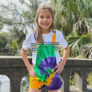 May include: A young person wearing a white t-shirt with green, purple, and gold stripes and tie-dye overalls. The overalls have a green, purple, and yellow tie-dye pattern and a green tassel.