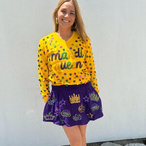 May include: A yellow long-sleeve shirt and a purple skirt. The shirt has the text "mardi queen" and is decorated with colorful sequins. The purple skirt is embroidered with crowns, stars, and masks. The outfit is festive and colorful.