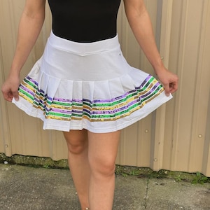 May include: White pleated skirt with a colorful sequin stripe around the hem. The sequins are in shades of green, gold, purple, and yellow.