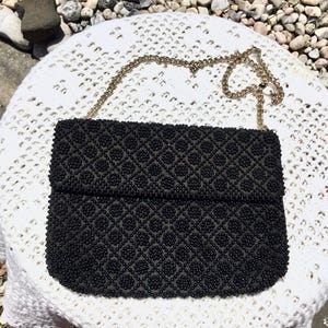 May include: A black beaded clutch bag with a gold chain strap. The bag has a geometric pattern and is made of small black beads.